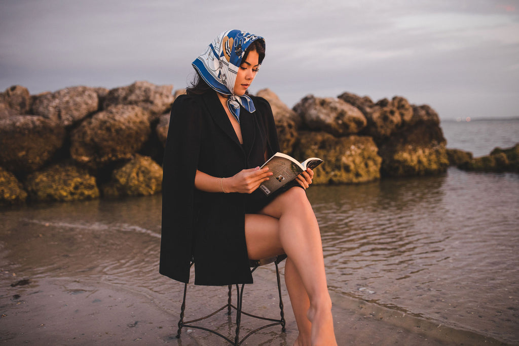 Woman wearing a Nina Nieves navy blue cape (Marina) at the beach reading a book. 